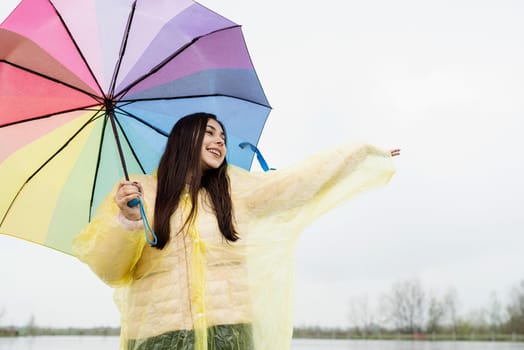 Beautiful smiling brunette woman in yellow raincoat holding rainbow umbrella out in the rain, catching raindrops with her hand