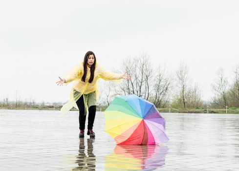 Beautiful brunette woman with funny face in yellow raincoat catching colorful umbrella outdoors in the rain. Rainbow umbrella lying on the ground