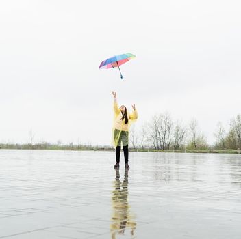 Beautiful brunette woman in yellow raincoat holding rainbow umbrella out in the rain