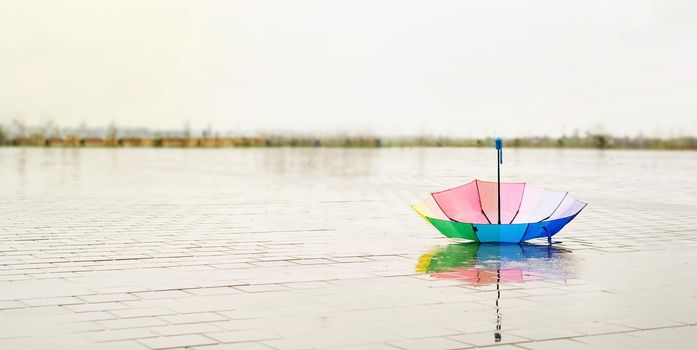 Unused colorful umbrella lying upside down on ground being rained upon. Rainbow colored umbrella lying in puddles on the wet street ground. Copy space