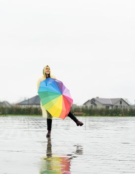 Beautiful brunette woman in yellow raincoat holding rainbow umbrella out in the rain