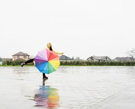 Beautiful brunette woman in yellow raincoat holding rainbow umbrella out in the rain