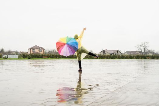 Beautiful brunette woman in yellow raincoat holding rainbow umbrella out in the rain