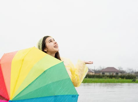 Beautiful smiling brunette woman in yellow raincoat holding rainbow umbrella out in the rain, catching raindrops with her hand