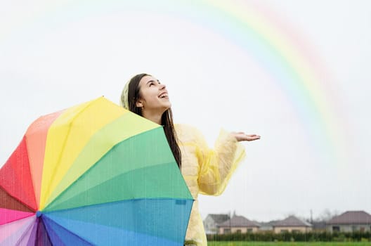 Beautiful smiling brunette woman in yellow raincoat holding rainbow umbrella out in the rain, catching raindrops with her hand