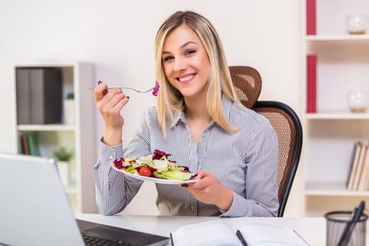 Beautiful businesswoman enjoys eating salad while working in her office.