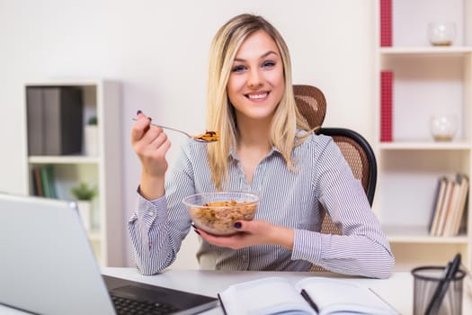 Beautiful businesswoman enjoys eating corn flakes for breakfast while working in her office.