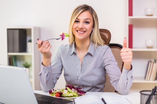Beautiful businesswoman enjoys eating salad and showing thumb up  while working in her office.