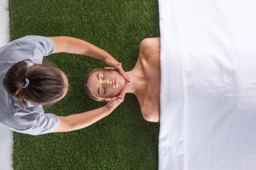 Facial massage, top view of beautiful young woman lying on back and keeping her eyes closed while massage therapist massaging her face