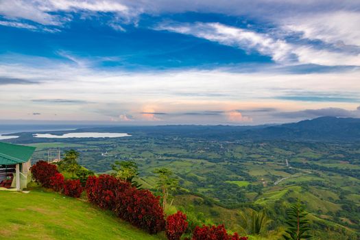View from a large mountain to a valley with a lake in the Dominican Republic. Setting sun. Around greenery and red bushes