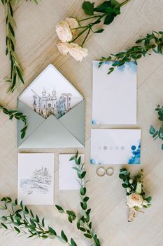 Wedding rings lie on the tile floor, surrounded by watercolor paintings, green twigs and flowers. The paintings depict old houses. High quality photo