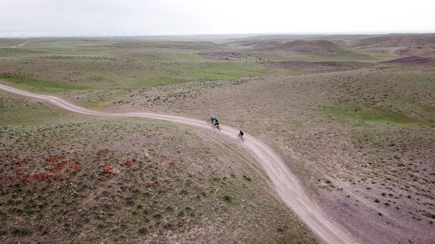 A group of cyclists rides along the windmills on a green field. Dusty road and cloudy weather at sunset. Blue clouds. In the distance, the Kapchagai Sea. Nature of Kazakhstan, Almaty. Renewable energy