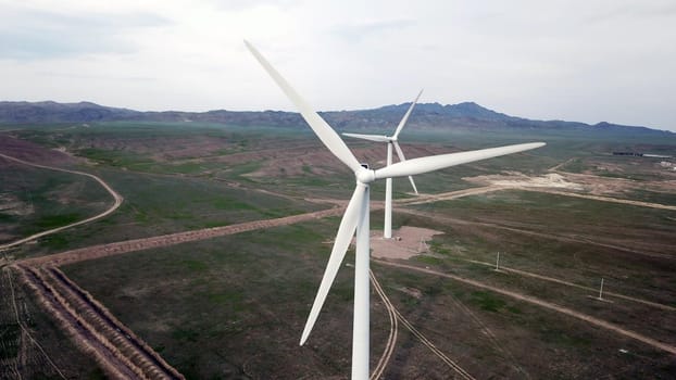 Windmills stand in the middle of green fields. Clean energy is generated. Cloudy weather, dark clouds. Green grass grows. High hills in the distance. Top view from drone. Industrial farm of Kazakhstan