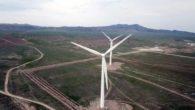 Windmills stand in the middle of green fields. Clean energy is generated. Cloudy weather, dark clouds. Green grass grows. High hills in the distance. Top view from drone. Industrial farm of Kazakhstan