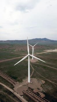 Windmills stand in the middle of green fields. Clean energy is generated. Cloudy weather, dark clouds. Green grass grows. High hills in the distance. Top view from drone. Industrial farm of Kazakhstan