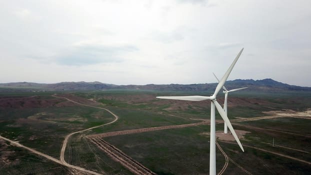 Windmills stand in the middle of green fields. Clean energy is generated. Cloudy weather, dark clouds. Green grass grows. High hills in the distance. Top view from drone. Industrial farm of Kazakhstan