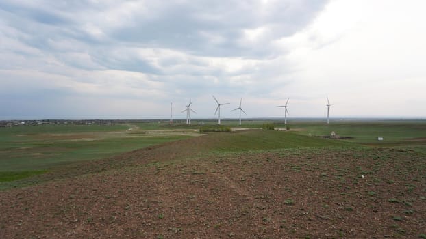 Windmills stand in the middle of green fields. Clean energy is generated. Cloudy weather, dark clouds. Green grass grows. High hills in the distance. Top view from drone. Industrial farm of Kazakhstan