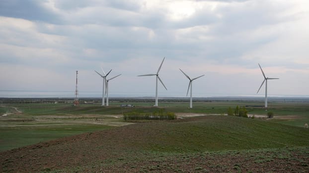 Windmills stand in the middle of green fields. Clean energy is generated. Cloudy weather, dark clouds. Green grass grows. High hills in the distance. Top view from drone. Industrial farm of Kazakhstan