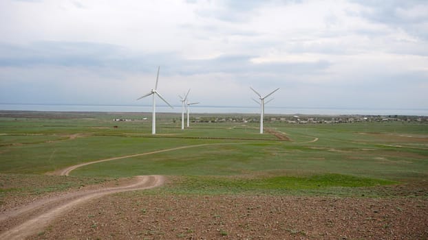 Windmills stand in the middle of green fields. Clean energy is generated. Cloudy weather, dark clouds. Green grass grows. High hills in the distance. Top view from drone. Industrial farm of Kazakhstan