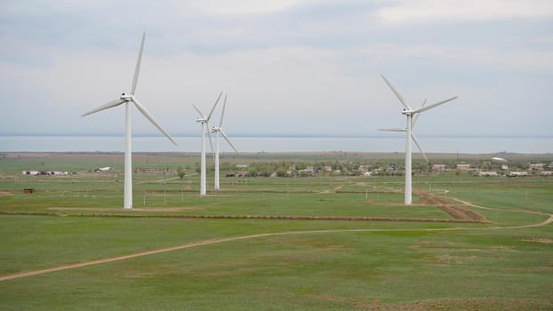 Windmills stand in the middle of green fields. Clean energy is generated. Cloudy weather, dark clouds. Green grass grows. High hills in the distance. Top view from drone. Industrial farm of Kazakhstan