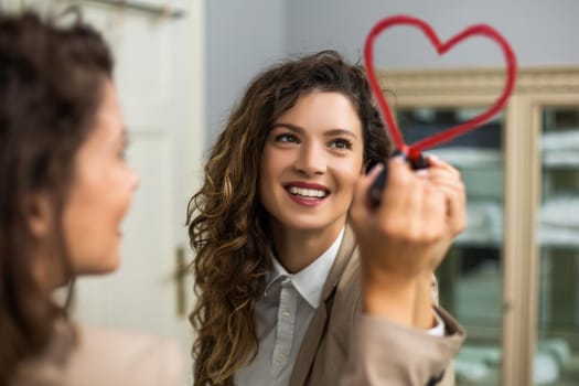 Businesswoman is drawing heart with lipstick on the mirror while preparing for work.
