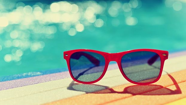 Summer background. Concept for summer and vacation. Red sunglasses by the pool in the background with blue water.