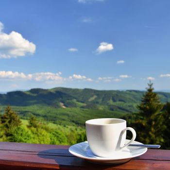Morning cup of coffee with a beautiful mountain landscape background. White cup and saucer - espresso on a wooden table.