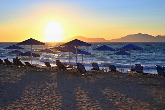 Sunbeds and umbrella on the beach at sunset by the sea. Beautiful concept for vacation, summer holidays and travel.
