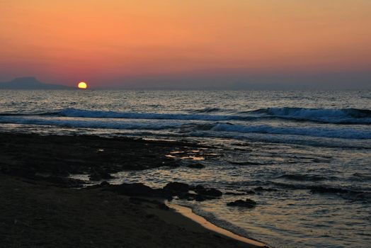 Beautiful summer sunset by the sea. Amazing scenery on the beach with waves and reflection of the sun. Background for holiday and vacation travel. Greece Crete Island.