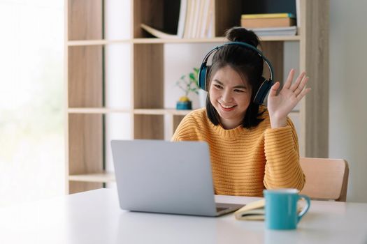 happy young asian ethnicity female manager wearing wireless headphones, looking at laptop screen, holding pleasant conversation with partners clients online, working remotely at workplace..