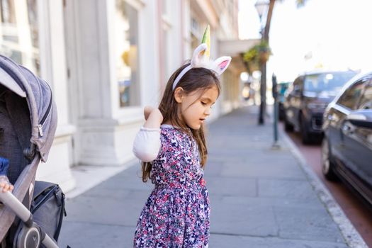 Adorable view of little girl wearing colorful dress and unicorn headband in the street. Portrait of cute young child with unicorn horn and ears standing on the sidewalk. Lovely kids outdoors