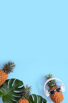 Funny pineapple wearing white headphone, concept of listening music, isolated on blue background with tropical palm leaves, top view, flat lay design.