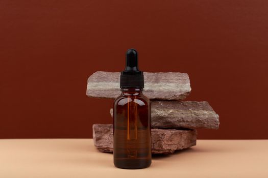 Selective focus, skin serum in dark glass bottle next to stack of stones on beige table against brown background with copy space. Concept of skin care products for young looking, glowing skin