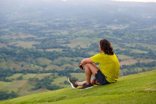 In summer, a man with long hair sits on the green slope of the mountain, looking into the distance at the valley. Dominican Republic, sunset in the mountains