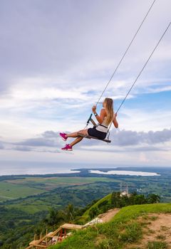 A young girl, blonde, swinging on a swing on a mountain slope in summer. Swing high in the mountains above the valley. Dominican Republic. Setting sun