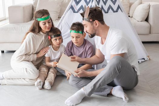 Happy family of parents and two children playing indian at home, wigwam tent, feather roach, reading book about native american indian culture