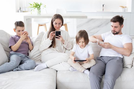 Caucasian parents and kids use devices together sit on sofa, tech addicted family with children hold phone digital tablet having fun with gadgets at home, technology dependence