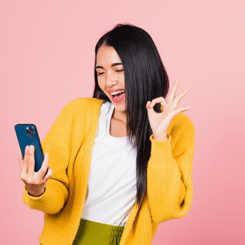 Happy Asian portrait beautiful cute young woman excited holding mobile phone and gesturing ok sign, studio shot isolated on pink background, Thai female making finger symbol on smartphone
