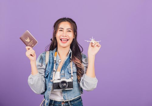 Happy Asian portrait beautiful young woman excited smiling in summer vacation holding airplane toy, passport and vintage photo camera, Thai female ready travel trip isolated on purple background
