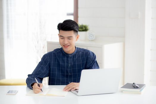 Young asian business man writing on notebook for planning working and using laptop computer on desk at home, notes about finance, male study and learning, business and communication concept.