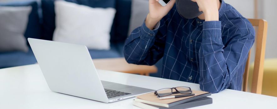 Young asian business man in face mask talking smartphone and work on laptop computer for protect covid-19, businessman quarantine work from home with notebook, social distancing and stay home.