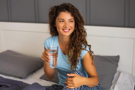 Beautiful woman drinking water while sitting on bed in her bedroom.