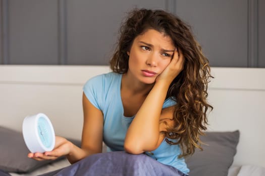 Sad woman in pajamas holding alarm clock in bed.