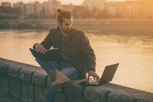 Handsome businessman using personal organizer and laptop while sitting by the river.
