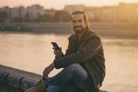 Handsome modern businessman using phone while sitting by the river.
