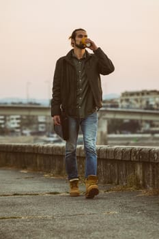 Handsome modern businessman with laptop bag drinking coffee and walking by the river.