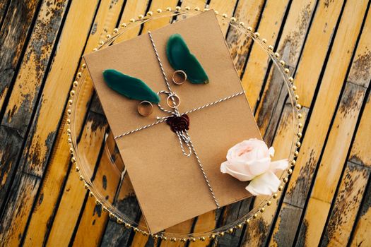 Beautiful brown envelope tied with a rope with rings, a rose and petals lies on a transparent tray on a wooden table. High quality photo