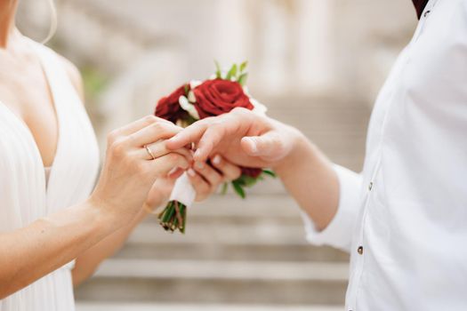 The bride puts a wedding ring on the groom's finger and holds a bouquet of red and white roses in her hand . High quality photo