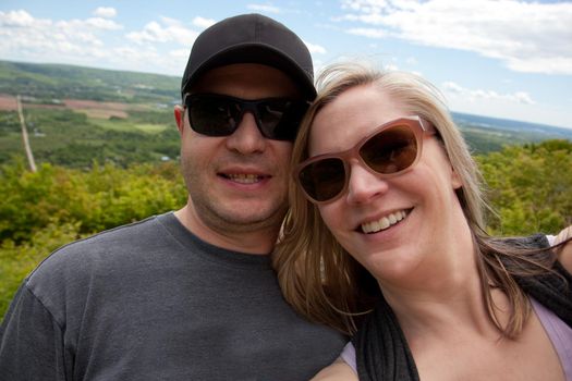  A smilng couple holds the camera or phone in front of them and takes a selfie by a beautiful summer valley 