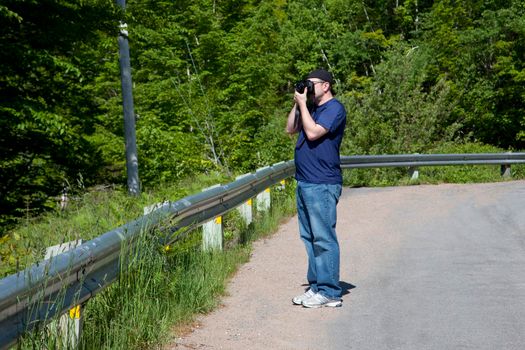  A man with a camera takes photos outside by leafy trees on a roadside